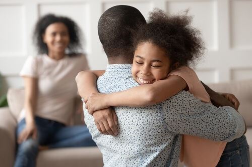 Smiling girl hugs her father while her smiling mother sits in the background.